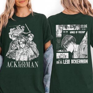 May include: Dark green t-shirt featuring a black and white graphic of anime characters. The front of the shirt displays the word "ACKERMAN" in white. The back of the shirt has a manga-style design with Japanese text and the words "WINGS OF FREEDOM".