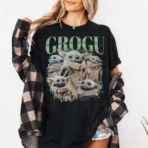 Puede incluir: Camiseta negra con la palabra "GROGU" en verde sobre un gr&aacute;fico de Grogu, tambi&eacute;n conocido como Baby Yoda, de la serie Star Wars. El gr&aacute;fico muestra a Grogu en varias poses. La camiseta es de cuello redondo.