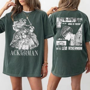 May include: Dark green t-shirt featuring a black and white graphic of anime characters with the text "ACKERMAN" on the front. The back of the shirt displays additional anime imagery and the text "LEVI ACKERMAN".