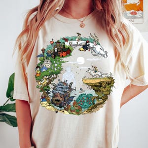 May include: A cream-coloured t-shirt with a circular graphic depicting animated characters and scenes. The design includes a white dragon, a girl on a broom, and various buildings and creatures in a colourful, detailed illustration.