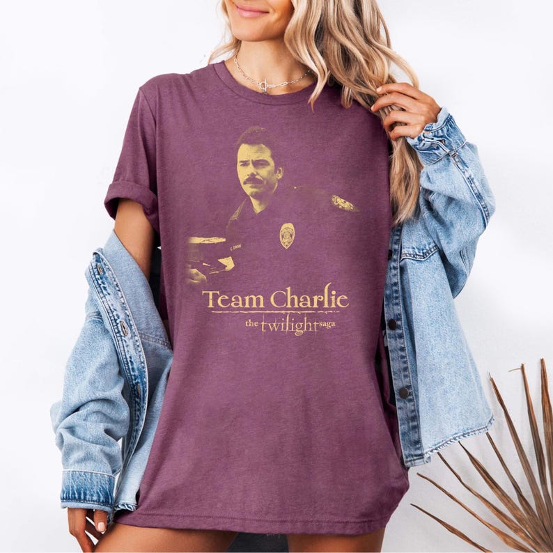 May include: A maroon t-shirt featuring a sepia-toned image of a man with a mustache and the text "Team Charlie" and "the twilight saga" in gold lettering. The shirt has short sleeves and a crew neck.