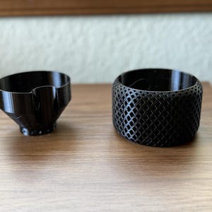 May include: Two black 3D-printed objects sit on a wooden surface. One is a small, angled container. The other is a cylindrical band with a diamond-patterned mesh design. The objects are likely decorative or functional.