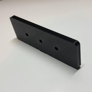 Puede incluir: Un objeto rectangular negro con tres agujeros espaciados. El objeto tiene una superficie texturizada y está en ángulo, con un borde ligeramente levantado. Parece estar hecho de un material sólido.