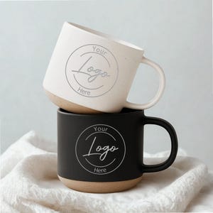 May include: Two ceramic mugs, one cream and one black, stacked. Both have a brown base and a curved handle. The cream mug features the text "Your Logo Here" within a circle. The black mug also has the same text.