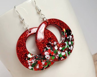 May include: Red and green glitter hoop earrings with silver hooks. The earrings are made of resin and have a sparkly finish.