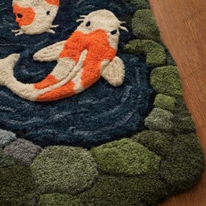 May include: A detailed rug featuring a koi fish design. The rug has a dark blue background with two koi fish in orange and white. Green and gray textured shapes represent plants and stones. The rug is made of a soft, textured material.