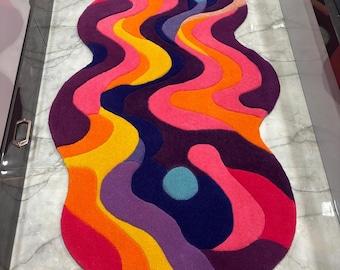 May include: An irregularly shaped area rug featuring a vibrant, multi-colored pattern of flowing, wavy lines in shades of hot pink, bright orange, sunny yellow, deep purple, and dark navy blue, accented by small, circular light blue spots.