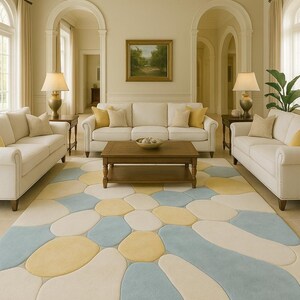May include: A living room with three white sofas, each adorned with yellow and beige pillows. A wooden coffee table sits in front of the center sofa. The room features a large, colorful rug with blue, yellow, and cream-colored pebble-like shapes.