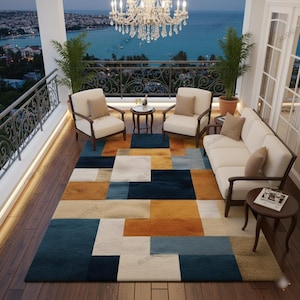 May include: A rectangular area rug featuring a geometric pattern of large, interlocking blocks in shades of navy blue, mustard yellow, cream, and light blue. The rug is positioned on a dark wood floor in a balcony setting with outdoor furniture.