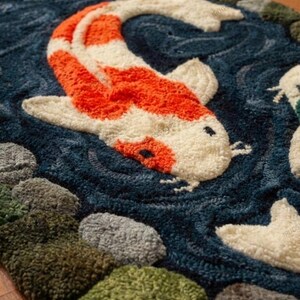 May include: A detailed rug featuring a koi fish design. The fish is orange and white, set against a dark blue water background. The rug has a textured appearance, with green, gray, and brown stones along the edges, creating a pond scene.