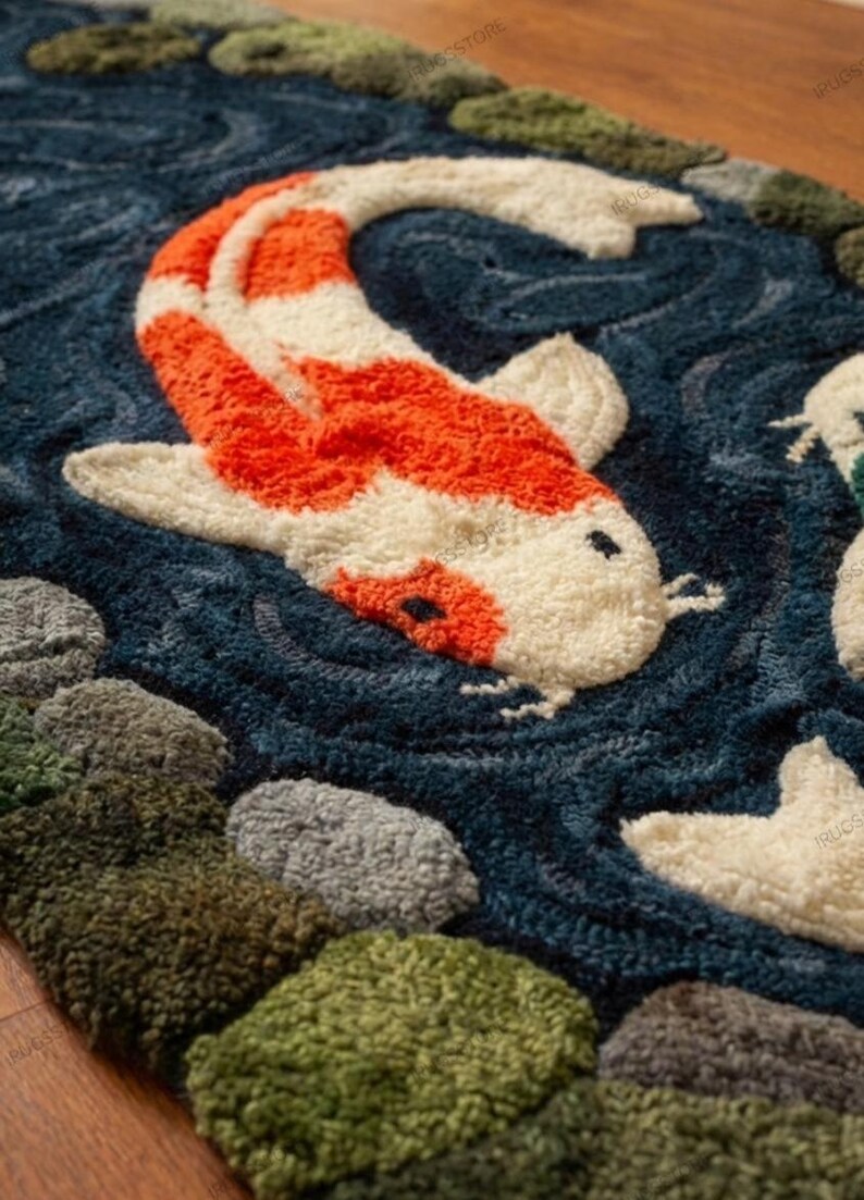 May include: A detailed rug featuring a koi fish design. The fish is orange and white, set against a dark blue water background. The rug has a textured appearance, with green, gray, and brown stones along the edges, creating a pond scene.