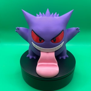 May include: A purple Gengar figurine with red eyes and a pink tongue, mounted on a black circular base. The figurine has a spiky head and outstretched arms, set against a green background.