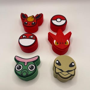 May include: A collection of colorful, character-shaped accessories. The assortment includes red, green, and tan figures with various designs, including a Pokeball and several stylized creatures. The items are arranged on a white surface.