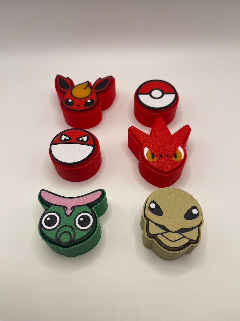 May include: A collection of colorful, character-shaped accessories. The assortment includes red, green, and tan figures with various designs, including a Pokeball and several stylized creatures. The items are arranged on a white surface.
