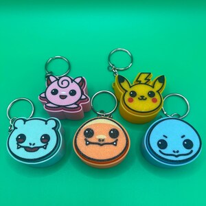 May include: Six colorful character keychains. The keychains feature a pink, yellow, blue, and orange design. Each keychain has a silver metal ring. The background is a solid green color.