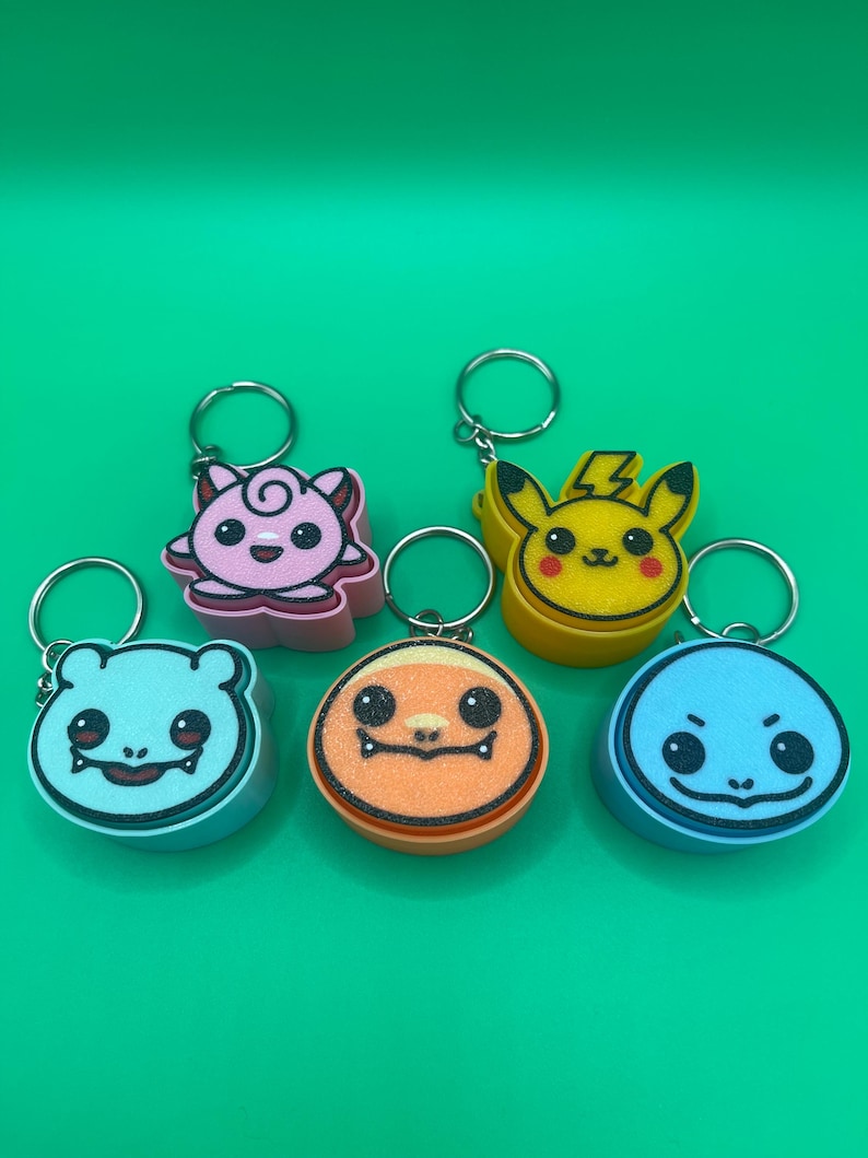 May include: Six colorful character keychains. The keychains feature a pink, yellow, blue, and orange design. Each keychain has a silver metal ring. The background is a solid green color.