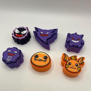 May include: A collection of colorful, cartoon-style character figures. The figures feature various designs in purple, orange, and black, with distinct facial expressions. The figures are arranged on a white surface.