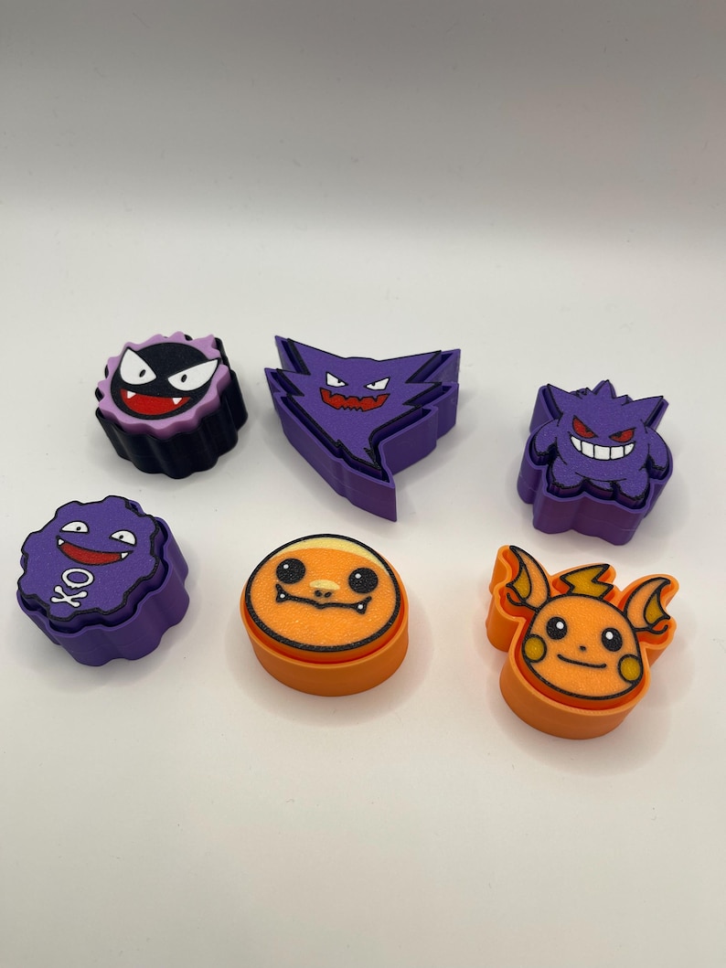 May include: A collection of colorful, cartoon-style character figures. The figures feature various designs in purple, orange, and black, with distinct facial expressions. The figures are arranged on a white surface.