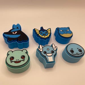 May include: Six colorful, cartoon character-shaped erasers. The erasers are in shades of blue, green, and yellow. Each eraser features a different character design, with detailed facial features and outlines.