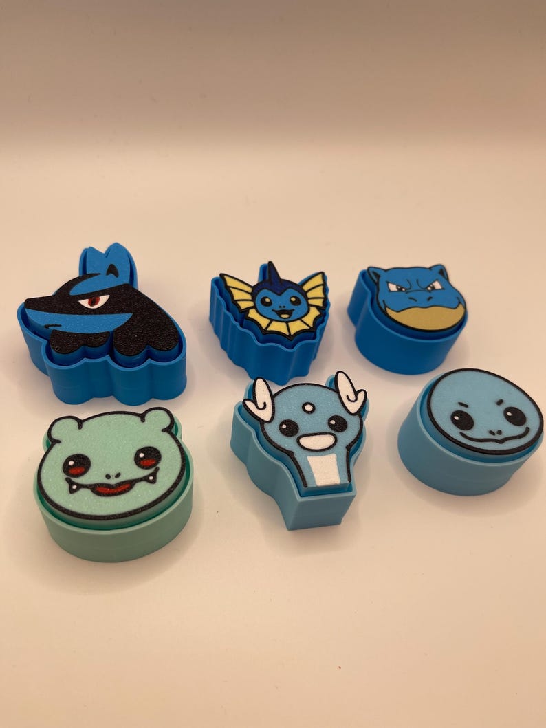 May include: Six colorful, cartoon character-shaped erasers. The erasers are in shades of blue, green, and yellow. Each eraser features a different character design, with detailed facial features and outlines.