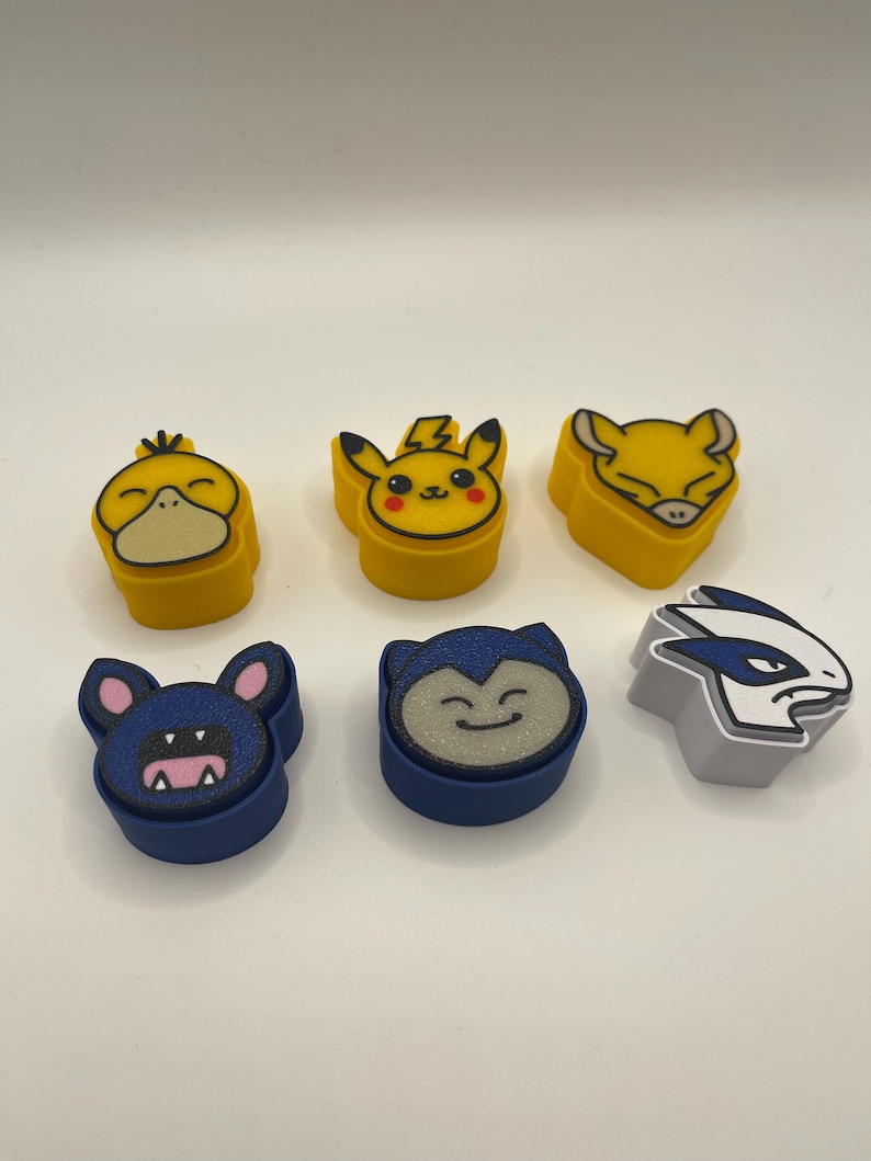 May include: Six colorful character-shaped erasers. The erasers feature designs of popular characters in yellow, blue, and gray. The characters include a duck, Pikachu, a fox, a bat, a Snorlax, and a Lugia.