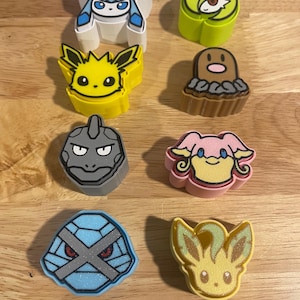May include: A collection of eight colorful character-shaped erasers. The erasers feature various designs, including a blue and white ice-themed character, a yellow electric-type character, and a gray rock-type character. The erasers are arranged on a light brown wooden surface.