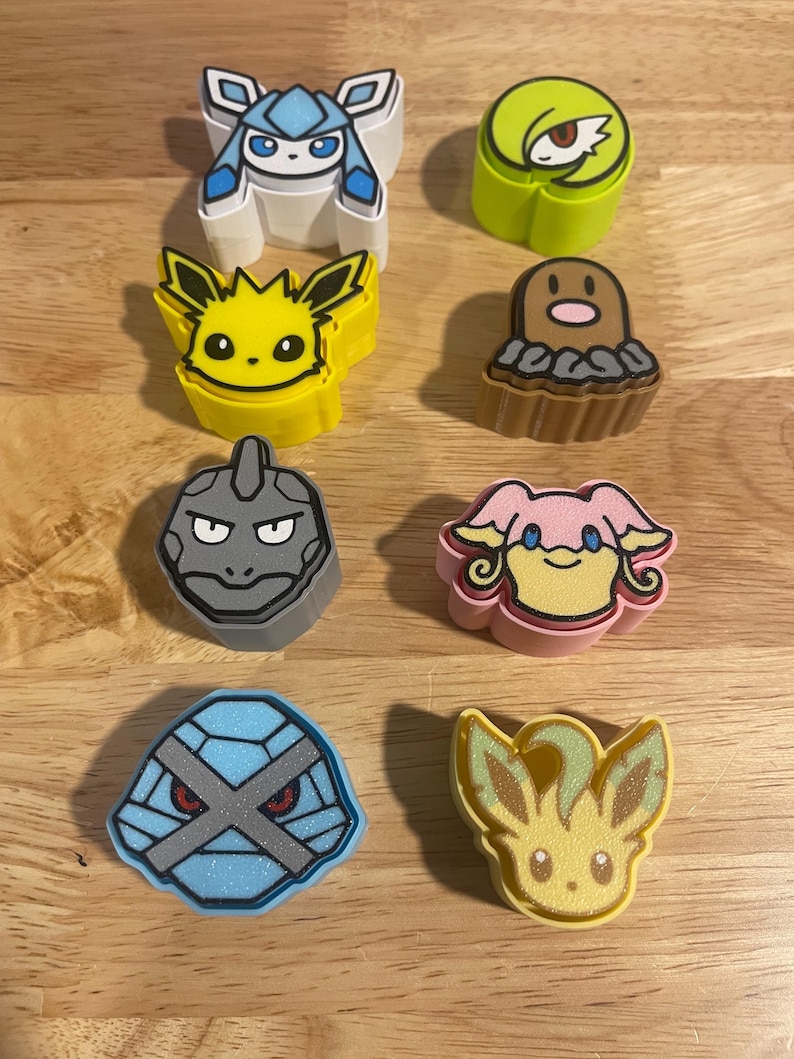 May include: A collection of eight colorful character-shaped erasers. The erasers feature various designs, including a blue and white ice-themed character, a yellow electric-type character, and a gray rock-type character. The erasers are arranged on a light brown wooden surface.