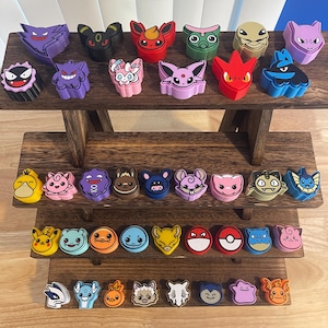 May include: A collection of colorful character-shaped figurines, including a purple Gengar, a red Flareon, and a blue Blastoise, displayed on a multi-tiered wooden stand. The figurines are arranged in rows, showcasing a variety of designs and colors.