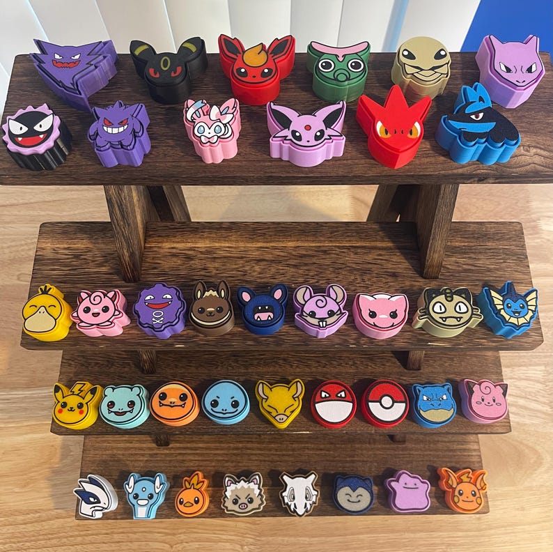 May include: A collection of colorful character-shaped figurines, including a purple Gengar, a red Flareon, and a blue Blastoise, displayed on a multi-tiered wooden stand. The figurines are arranged in rows, showcasing a variety of designs and colors.