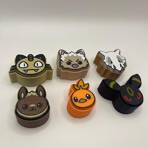 May include: Six colorful, cartoon character-shaped charms. The charms feature various designs, including a cat, a hedgehog, a skull, a rabbit, a chick, and a black creature with yellow accents. The charms are likely made of a rubbery material.