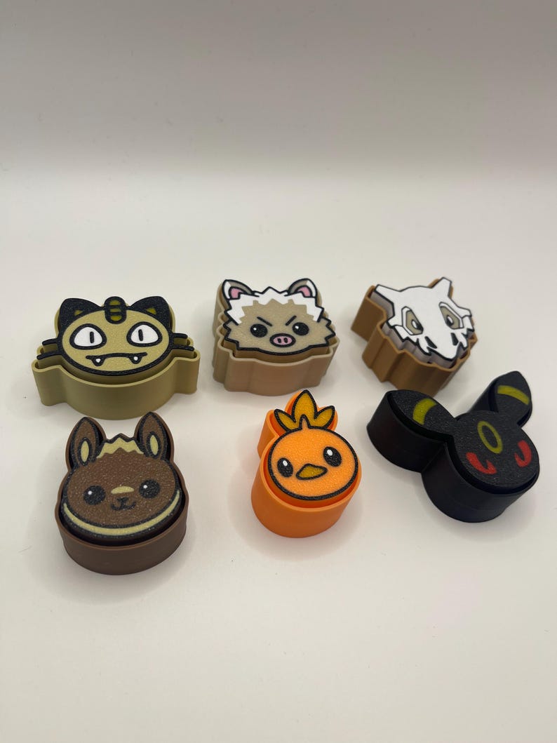 May include: Six colorful, cartoon character-shaped charms. The charms feature various designs, including a cat, a hedgehog, a skull, a rabbit, a chick, and a black creature with yellow accents. The charms are likely made of a rubbery material.