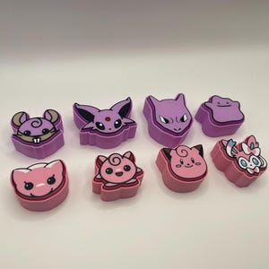 May include: A collection of colorful, cartoon-style character figures in shades of pink and purple. The figures feature various animal-like designs with distinct facial features and shapes. The figures are arranged in two rows on a white surface.