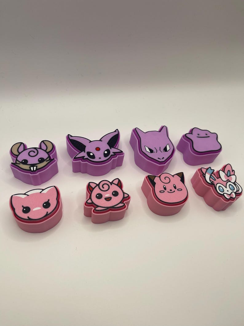 May include: A collection of colorful, cartoon-style character figures in shades of pink and purple. The figures feature various animal-like designs with distinct facial features and shapes. The figures are arranged in two rows on a white surface.