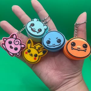 May include: A collection of colorful character keychains featuring cartoon designs. The keychains include a pink creature, a yellow Pikachu, a light blue creature, a blue character, and an orange character. Each keychain has a silver metal ring.