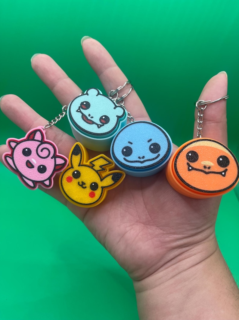 May include: A collection of colorful character keychains featuring cartoon designs. The keychains include a pink creature, a yellow Pikachu, a light blue creature, a blue character, and an orange character. Each keychain has a silver metal ring.