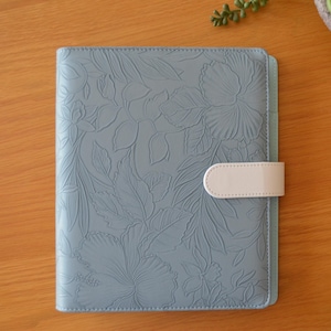 May include: A pale blue journal with a floral embossed design and a cream clasp. The cover has a detailed, raised pattern of leaves and flowers. The journal is closed and rests on a wooden surface.