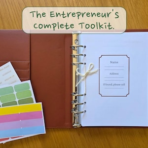 May include: A brown leather binder with the text "The Entrepreneur's complete Toolkit." Inside are lined pages, a pen, and stickers. The binder is open, revealing a section for contact information and a pen.