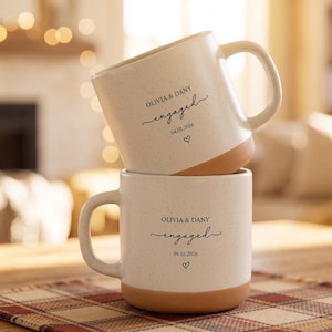 Puede incluir: Dos tazas de cerámica apiladas con un acabado blanco moteado y una base marrón. Cada taza tiene un asa y las palabras "OLIVIA & DANY engaged 04.01.2026" con un corazón. Las tazas están sobre un mantel individual a cuadros.