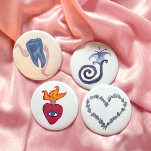 Puede incluir: Cuatro botones redondos blancos con diseños únicos de acuarela. Uno presenta un diente azul, otro una flor azul, un tercero un corazón rojo con una llama y el último un corazón morado hecho de hojas. Los botones están sobre una superficie de satén rosa.