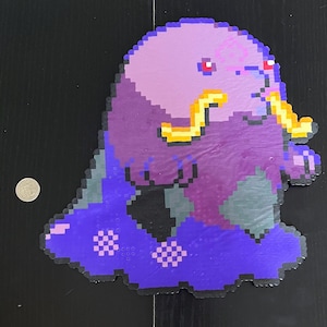 Può includere: Una figura di pixel art nei toni del viola, blu e giallo, che assomiglia a una creatura dei cartoni animati. La figura ha un corpo arrotondato, una testa grande e appendici gialle. I pixel rossi formano gli occhi. Una moneta è accanto alla figura per la scala.