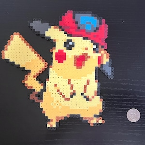 Pode incluir: Uma figura pixelizada de Pikachu feita de contas coloridas. A figura é amarela com bochechas vermelhas, um chapéu vermelho e uma viseira cinza e azul. A figura está segurando um objeto cinza. Há uma moeda ao lado da figura.