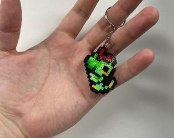 May include: A pixelated keychain featuring a green and black creature with red accents. The keychain is made of small, colorful beads and attached to a silver keyring. The design is reminiscent of retro video game characters.