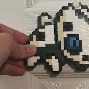 May include: Pixel art decoration featuring a white, black, and tan character with a blue eye. The design is made of small, colorful beads, and is attached to a white door. The character has a rounded shape.