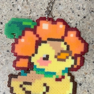 May include: A pixel art keychain featuring a yellow duckling with orange hair, a green leaf accessory, and a teal collar. The duckling has a small brown wing, pink cheeks, and orange feet. The design is constructed from small fused plastic beads.