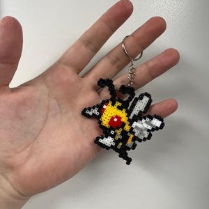 May include: A pixelated keychain featuring a bee design. The bee is primarily yellow, black, and white, with red accents. The keychain is attached to a silver metal ring.