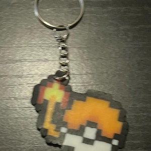 May include: Pixelated keychain featuring a torch design in shades of orange, yellow, and white, outlined in black. The keychain includes a silver key ring and chain, perfect for keys or bags.