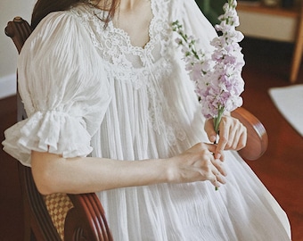 May include: A white nightdress with lace trim on the neckline and sleeves. The nightdress has a V-neck and ruffled sleeves. A person is holding a bunch of purple flowers while seated in a wooden chair. The style is vintage.