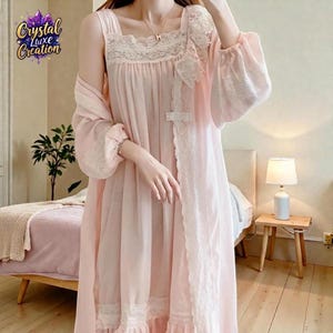 Pode incluir: Um conjunto de camisola e roupão rosa pálido com delicados detalhes em renda branca ao longo do decote quadrado, punhos e painel frontal. A peça possui mangas longas e largas, com uma silhueta fluida que vai até o chão com bainha.