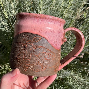 May include: Handmade ceramic mug with a pink glazed top and a brown base. The brown base features an embossed moth design. The mug has a curved handle and is held against a backdrop of green foliage.