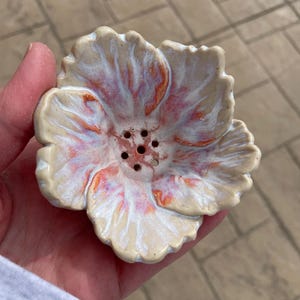 May include: A ceramic flower-shaped soap dish with a cream base and red and white accents. The dish has six drainage holes in the centre. The petals have a textured appearance. The dish is held in a hand.